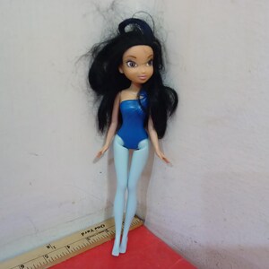 May include: A doll with black hair and a blue swimsuit. The doll has light blue legs and arms. The doll is standing on a red surface. A ruler is in the foreground.