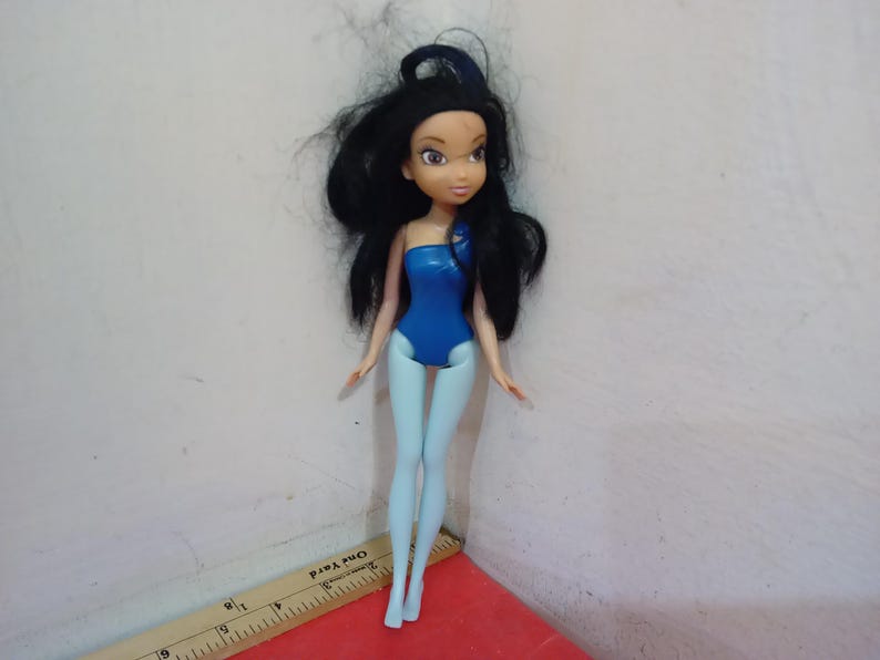 May include: A doll with black hair and a blue swimsuit. The doll has light blue legs and arms. The doll is standing on a red surface. A ruler is in the foreground.