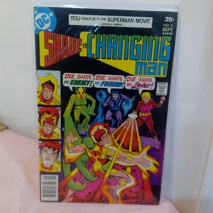 Puede incluir: Una portada de cómic con un superhéroe llamado "Shade the Changing Man". La portada es roja, amarilla y azul con un gráfico del superhéroe con un traje rojo y amarillo. El texto en la portada dice "You could be in the Superman Movie" y "Die, Shade, My Enemy! Die, Shade, My Friend! Die, Shade, My Lover!"