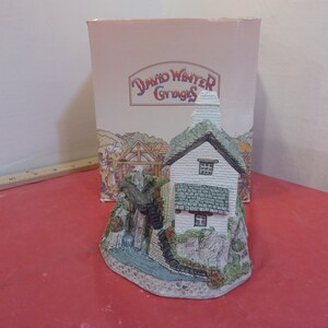 May include: A white cottage with a green roof and a stone chimney. The cottage is surrounded by a small stream and a waterfall. The cottage is part of a David Winter Cottages collection.