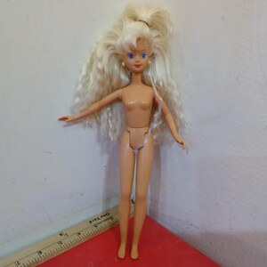 May include: A vintage doll with long, wavy blonde hair pulled back in a high ponytail. The doll has blue eyes and is wearing no clothing. The doll is standing with arms outstretched.