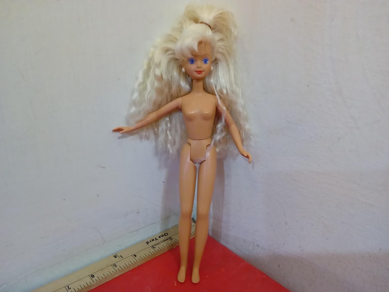 May include: A vintage doll with long, wavy blonde hair pulled back in a high ponytail. The doll has blue eyes and is wearing no clothing. The doll is standing with arms outstretched.
