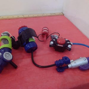 May include: A set of four toy water guns, including a green and blue gun with a clear tank, a blue and black gun with a black strap, a red and black gun with a red button, and a silver and purple gun with a blue handle.