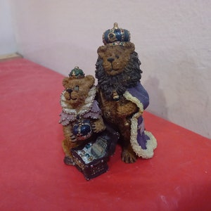 Puede incluir: Dos figuritas de peluche de osos vestidos de reyes, uno marrón y otro negro, con túnicas y coronas moradas y doradas. El oso marrón sostiene un cofre del tesoro.