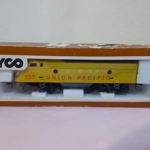 Könnte beinhalten: Eine gelbe Spielzeugeisenbahnlokomotive mit schwarzen Rädern und einem Tyco-Logo auf der Schachtel. Die Lokomotive trägt die Aufschrift "537 - UNION PACIFIC" an der Seite.