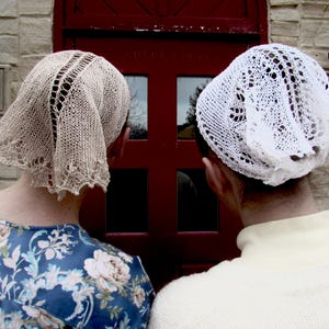 May include: Two women wearing knitted head coverings stand before a red door. One head covering is beige with a decorative openwork design, and the other is white with a lace pattern. The woman on the left wears a blue floral top.