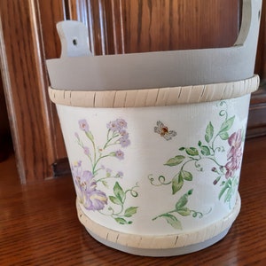 Op de afbeelding: Een witte houten plantenbak met een grijze rand en een bloemenontwerp met paarse, roze en groene bloemen. De plantenbak heeft een geweven strokenband rond de bovenkant.