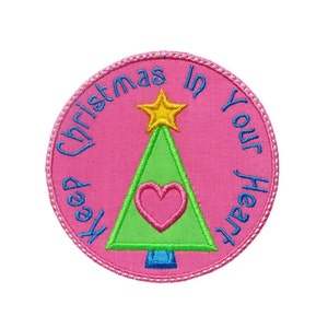 Pode incluir: Um patch redondo bordado rosa com o texto "Keep Christmas in Your Heart" em azul. Uma árvore de Natal verde com um coração rosa e uma estrela amarela está no centro. O patch tem uma borda costurada.