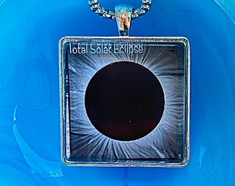May include: A silver chain necklace with a square pendant featuring a black and white photograph of a total solar eclipse. The pendant has the text "Total Solar Eclipse" printed on it.
