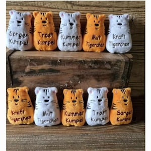 Könnte beinhalten: Zehn Plüschtiger mit gestickten Gesichtern und verschiedenen Sprüchen. Die Sprüche lauten: "Sorgen Kumpel", "Trost Kumpel", "Kummer Kumpel", "Mut Tigerchen", "Kraft Tigerchen", "Kraft Tigerchen", "Mut Tigerchen", "Kummer Kumpel", "Trost Kumpel" und "Sorge Kumpel".