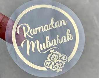 May include: A clear round sticker with a gold circle and the text "Ramadan Mubarak" in a gold cursive font. The sticker also features a gold decorative design.