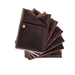 May include: A stack of brown leather wallets with different colored stitching. The top wallet has a brass button.