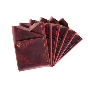 May include: A set of eight leather wallets in various shades of red, brown, and blue. Each wallet has a stitched edge and a small pocket on the front. The wallets are arranged in a fan shape, with the front of each wallet facing the viewer.