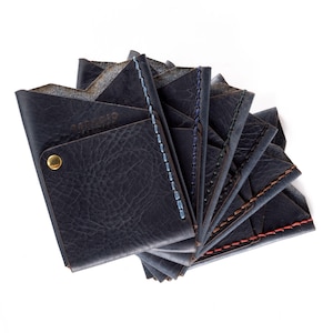 May include: A set of eight leather card holders in various shades of blue, brown, and black. Each holder features a stitched edge and the text "Espacio Handmade" embossed on the front.