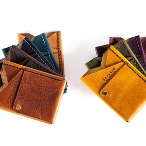May include: Two leather wallets in shades of brown, blue, and yellow are displayed against a white background. The wallets are open, showcasing multiple card slots. One wallet has a brass snap closure. The text "ESPACIO HANDMADE" is visible on one wallet.