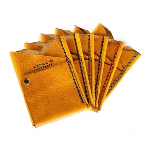 May include: A stack of eight yellow leather wallets with different colored stitching. The top wallet has the text "ESPACIO HANDMADE" embossed on the front.