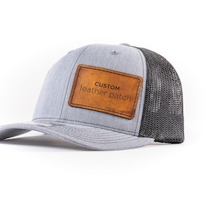 Può includere: Un cappello da camionista grigio e nero con una toppa in pelle marrone che dice "CUSTOM leather patch".