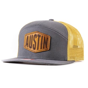 May include: A gray and gold trucker hat with a brown leather patch that says "AUSTIN" on it.