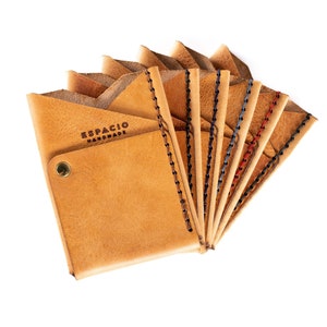 May include: A set of eight tan leather wallets with different colored stitching. The wallets are folded and fanned out, revealing the stitching detail. The wallets have the text "ESPACIO HANDMADE" embossed on the front.