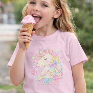 May include: A light pink t-shirt featuring a colorful unicorn design with a rainbow mane, horn, and decorative elements. The girl in the image is eating a pink ice cream cone.