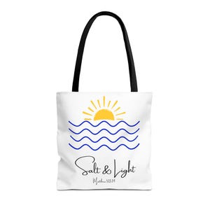 May include: White tote bag with black handles featuring a yellow sun over blue waves. The words "Salt & Light" are written in a script font, with "Matthew 5:13-14" below. The bag is designed for carrying items.