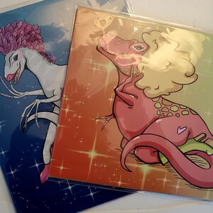 May include: Two rectangular art prints in clear protective sleeves. The left print features a white dinosaur with pink hair on a blue background with stars. The right print shows a pink dinosaur with blonde hair on an orange background with stars.