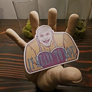 May include: A sticker featuring a smiling, bald character with a yellow outfit and a purple heart-shaped design. The text "IT MEANS TO" is visible on the sticker. The sticker is held by a wooden hand.