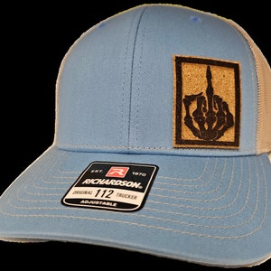 Op de afbeelding: Een lichtblauwe trucker-pet met een bruine patch met een zwarte skelethand die de middelvinger opsteekt. De pet heeft een wit mesh-achterpaneel en een zwart label met de tekst "Richardson. Est. 1970 Original 112 Trucker Adjustable".