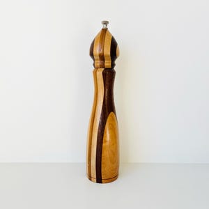 Puede incluir: Un molinillo de pimienta de madera con un diseño a rayas. La parte superior y el cuerpo del molinillo están hechos de franjas alternas de madera clara y oscura. La parte superior tiene una tapa de metal plateado.