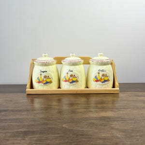 May include: A set of three cream-coloured ceramic canisters for sugar, tea, and coffee, each with a lid and a fruit and floral design. The canisters sit in a wooden rack.