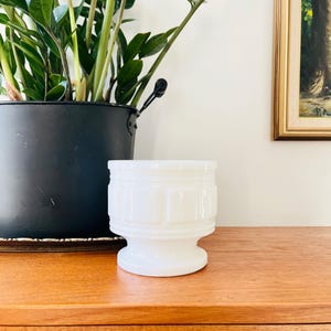 May include: White ceramic planter with a fluted design and a pedestal base. The planter is sitting on a wooden surface.