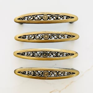 May include: Four antique brass cabinet pulls with intricate floral and scrollwork designs. Each pull is oval-shaped with a central flower and openwork detailing. The pulls are arranged in a vertical stack against a white marble background.