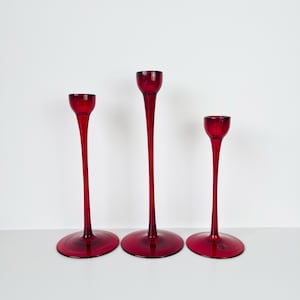 May include: Three red glass candle holders of varying heights. Each has a round base, a long, slender stem, and a small cup at the top. The candle holders are arranged in a row against a white background.