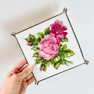 May include: Square decorative tray with a floral rose design. The tray features a white background with pink roses and green leaves. The tray has a silver metal frame.