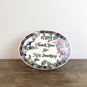 May include: An oval-shaped sign with a cream-coloured background featuring the black text Thank You for Not Smoking. The border is decorated with a colourful illustration of a hummingbird and pink fuchsia flowers with green leaves on a dark vine.