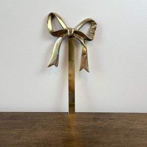 May include: A gold-coloured metal bow-shaped decorative object. The bow has a detailed design with flowing ribbon-like elements and is attached to a vertical bar. The object is likely a decorative accent or ornament.