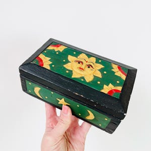 Könnte beinhalten: Eine rechteckige Holzkiste mit schwarzem Rahmen und grünem Deckel und Seiten. Der Deckel zeigt ein geschnitztes Sonnendesign mit roten Akzenten. Die Seiten haben Halbmonde und Sterne. Die Box wird von einer Hand gehalten.