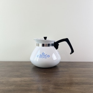 May include: A white teapot with a rounded body featuring a blue floral pattern. The teapot has a black handle, a black lid knob, and a silver-colored metal band around the top rim. The design includes blue flowers and leaves on the front surface.