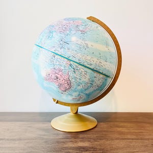 May include: A vintage globe featuring a light blue and pink map, supported by a beige stand and a wooden frame. This classic educational tool is suitable for home decor or educational use. The globe is approximately 30 cm tall.