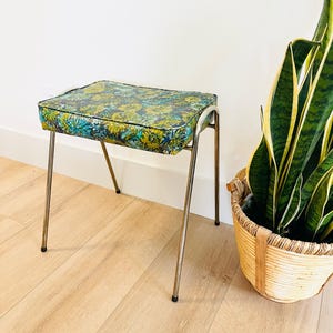 Puede incluir: Un taburete vintage con un asiento rectangular con un estampado floral en tonos verdes, azules y amarillos. Tiene patas cromadas. Una cesta de mimbre con una planta está al lado del taburete.