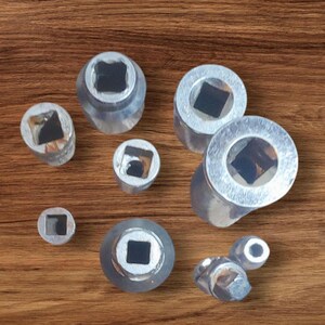 May include: A set of eight silver-colored metal sockets with square openings. The sockets are of varying sizes and are arranged on a wooden surface.