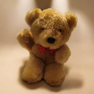 May include: A plush teddy bear in a light brown color, with a red bow tie featuring white polka dots. The bear has a black nose and is posed with one arm raised, giving a friendly appearance. A classic children's toy.