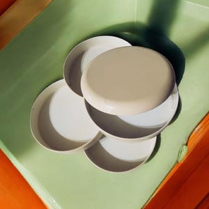 May include: A stack of small, white, round plates with a beige lid. The plates are arranged on a light green surface, with a shadow cast across them. The background includes orange and yellow surfaces.