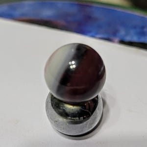 May include: A glass marble with a white, black, and maroon swirl pattern sits atop a silver metal ball bearing. The marble is round and reflective, with a glossy finish. The metal ball bearing is also round and has a shiny surface.