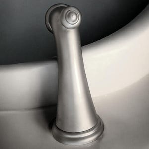 Pu&ograve; includere: Una maniglia del rubinetto in nichel spazzolato con una parte superiore decorativa e una base svasata. La maniglia &egrave; curva e montata su una superficie bianca, con uno sfondo grigio scuro.