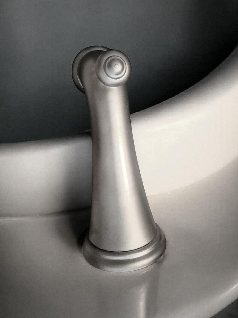 Pu&ograve; includere: Una maniglia del rubinetto in nichel spazzolato con una parte superiore decorativa e una base svasata. La maniglia &egrave; curva e montata su una superficie bianca, con uno sfondo grigio scuro.