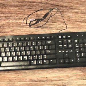 May include: A black computer keyboard with a standard QWERTY layout, a numeric keypad, and a thin black wired cable extending from the top. The keyboard features white lettering on each key and includes a dedicated Windows key on the bottom row.