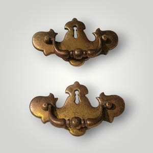 May include: Two antique brass drawer pulls. Each pull features a decorative design with a keyhole and a curved handle. The pulls have a warm, aged patina, suggesting vintage or retro style. Perfect for furniture restoration or adding a touch of classic charm.