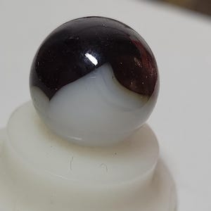 May include: A glossy, spherical marble with a striking contrast of colors. The top half is a deep, rich brown, transitioning to a bright white on the bottom. The marble is resting on a white, rounded base.