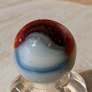 May include: A spherical glass marble featuring a swirling pattern of opaque white, translucent blue, and deep reddish-brown colors. The surface is smooth and reflective, showcasing a distinct blend of these three colors that wrap around the orb.